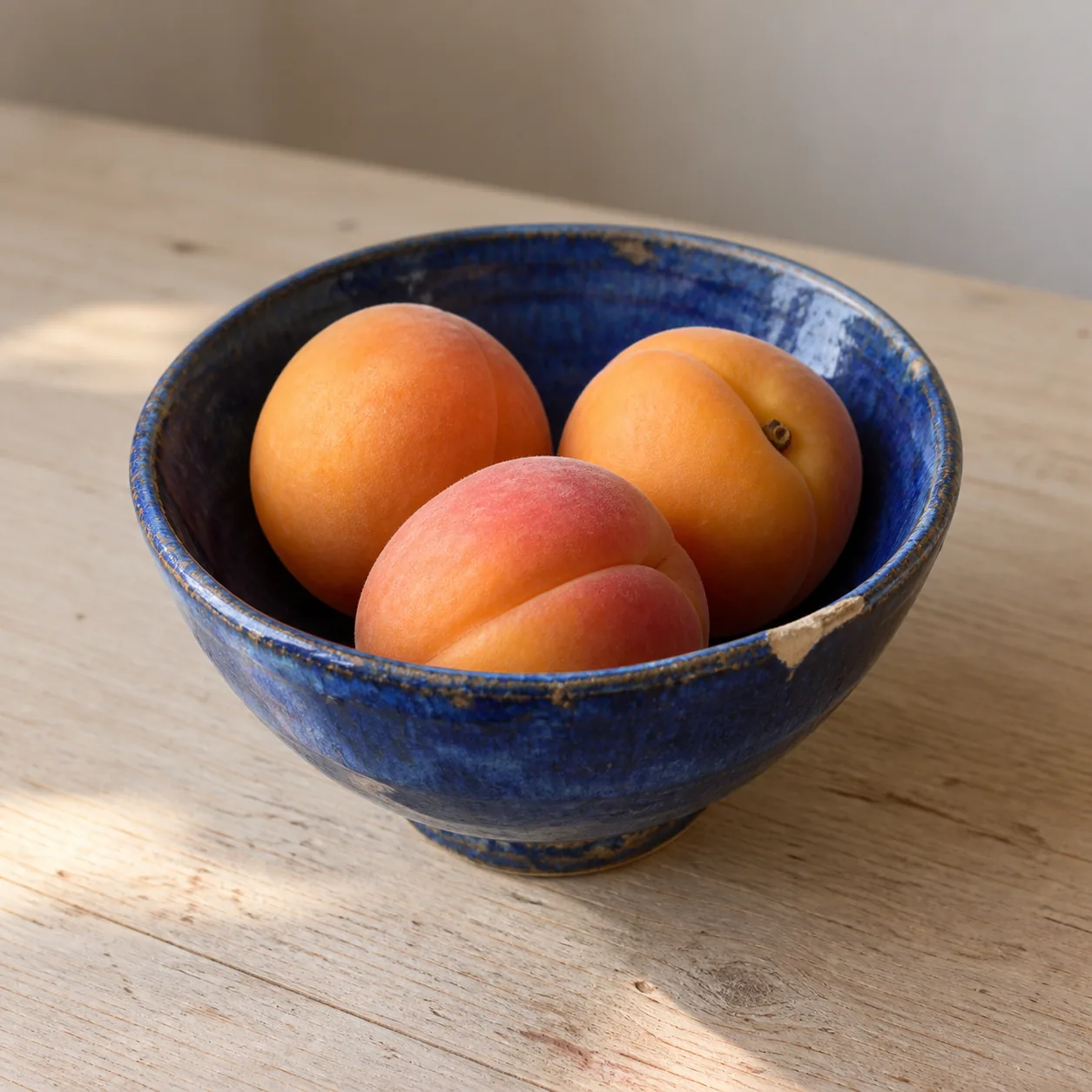 A chipped blue ceramic bowl with five apricots in gentle morning light