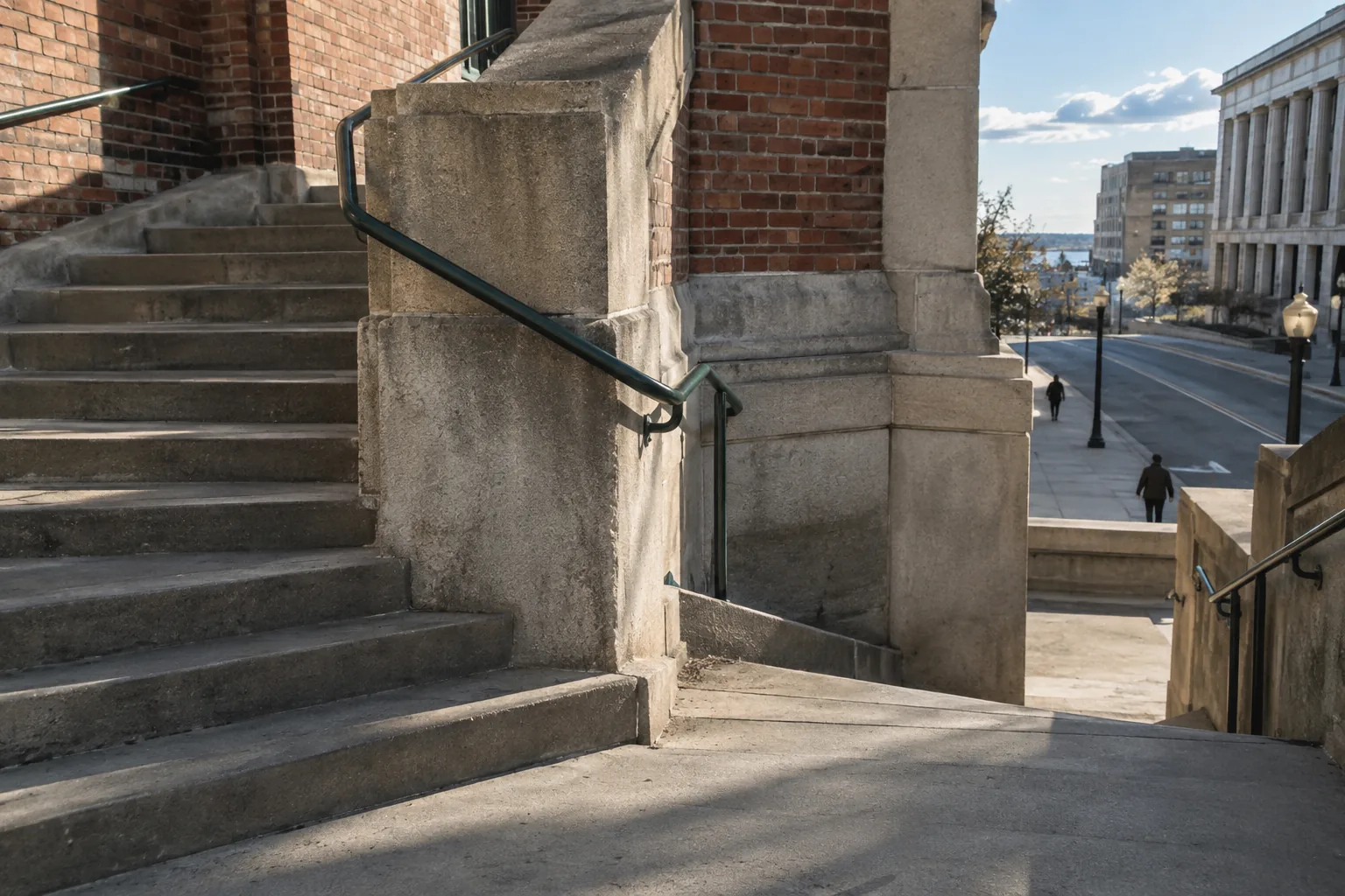 A public staircase turns at a sunlit landing, opening toward the sky and street beyond.