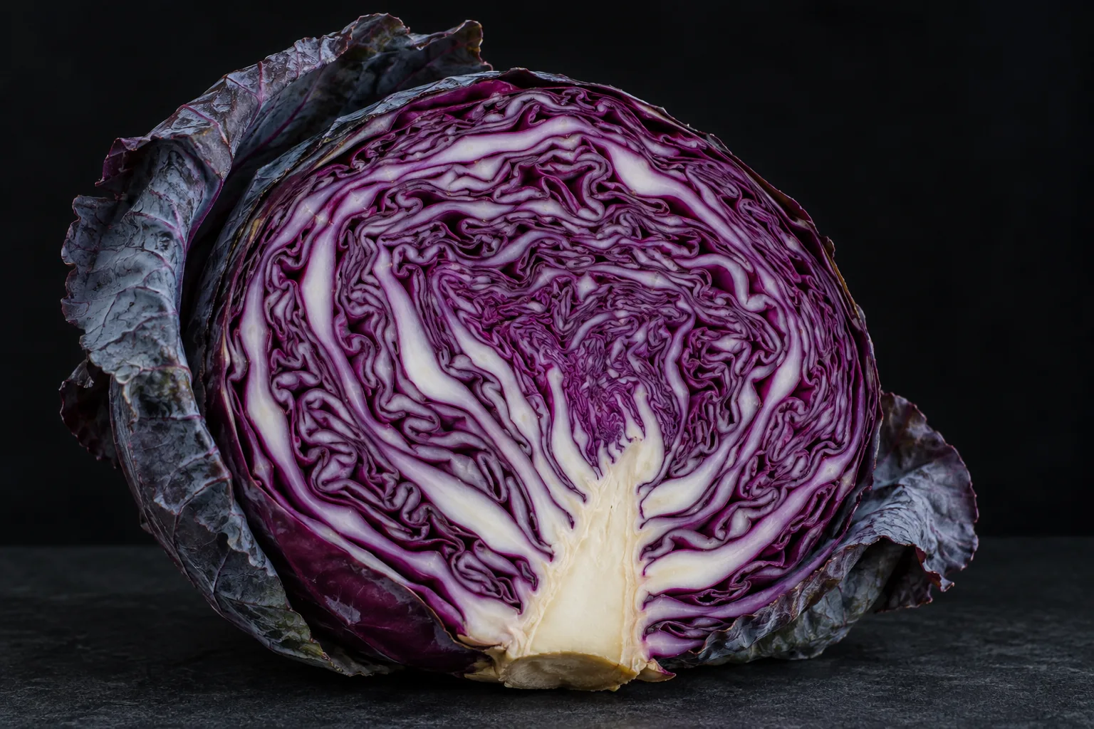 A halved red cabbage shows pale branching ribs among tightly folded violet leaves against a dark background.